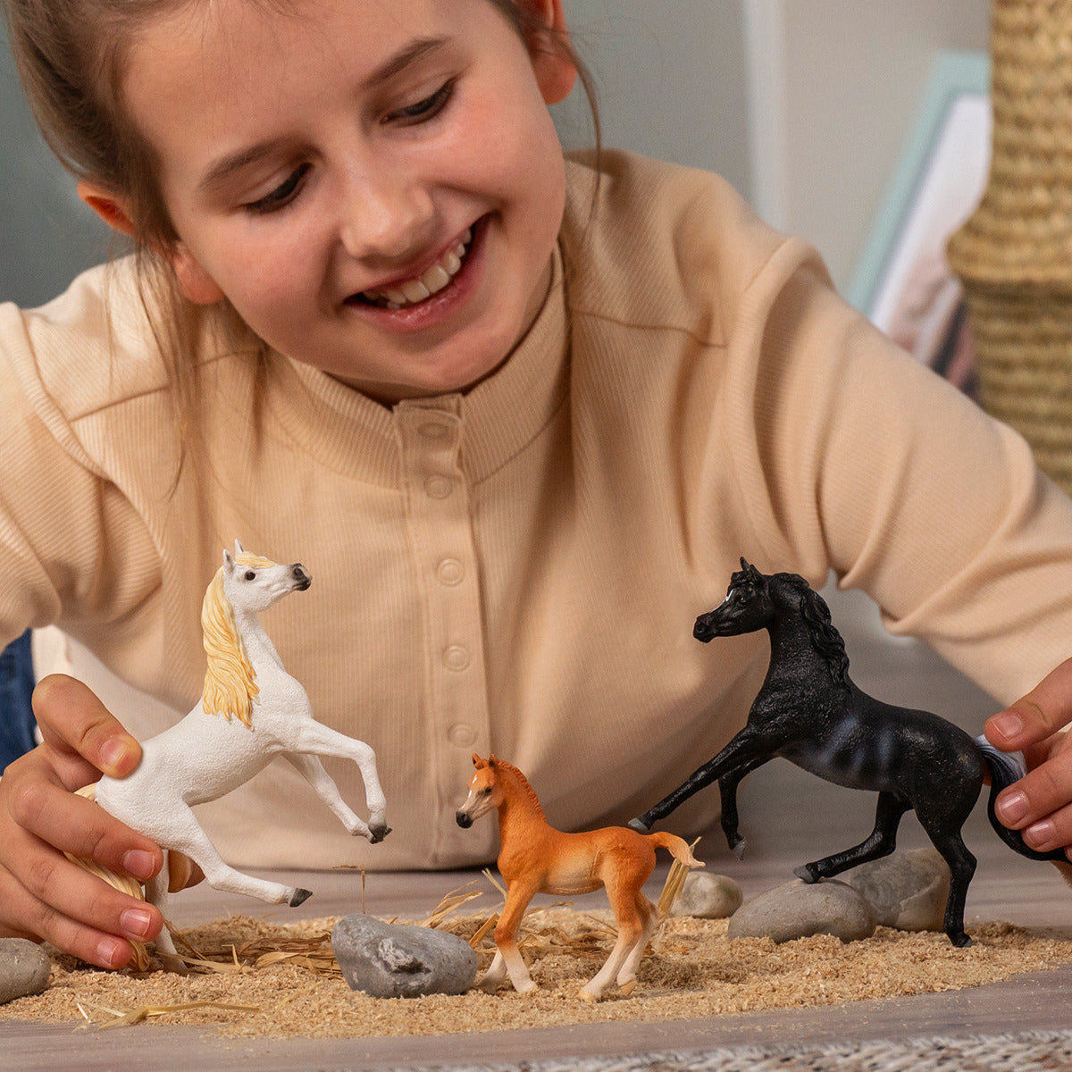 Smiling girl plays with three horse figurines (white, black, and foal) on a miniature sandy landscape