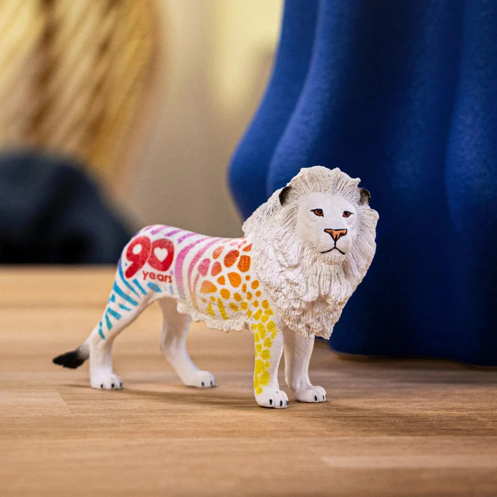 A white lion toy with rainbow patterns and red "90 years" text, standing on a wooden surface