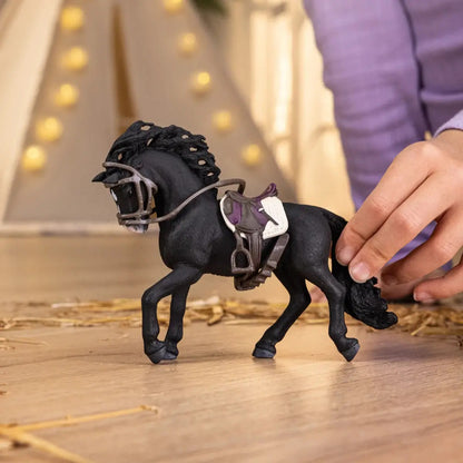 A child's hand touches a black toy horse figurine, saddled and bridled, on a wooden floor with straw