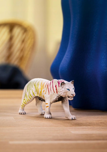 A white toy tiger with rainbow stripes and blue eyes walks on a wooden surface