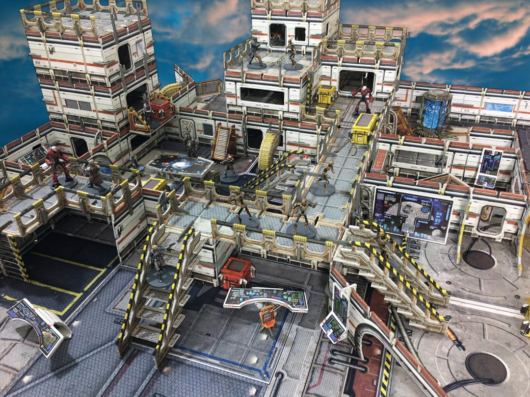 Sci-fi tabletop game: Miniature figures are arrayed across a modular, multi-level futuristic base with stairs and consoles, under a cloudy sky.