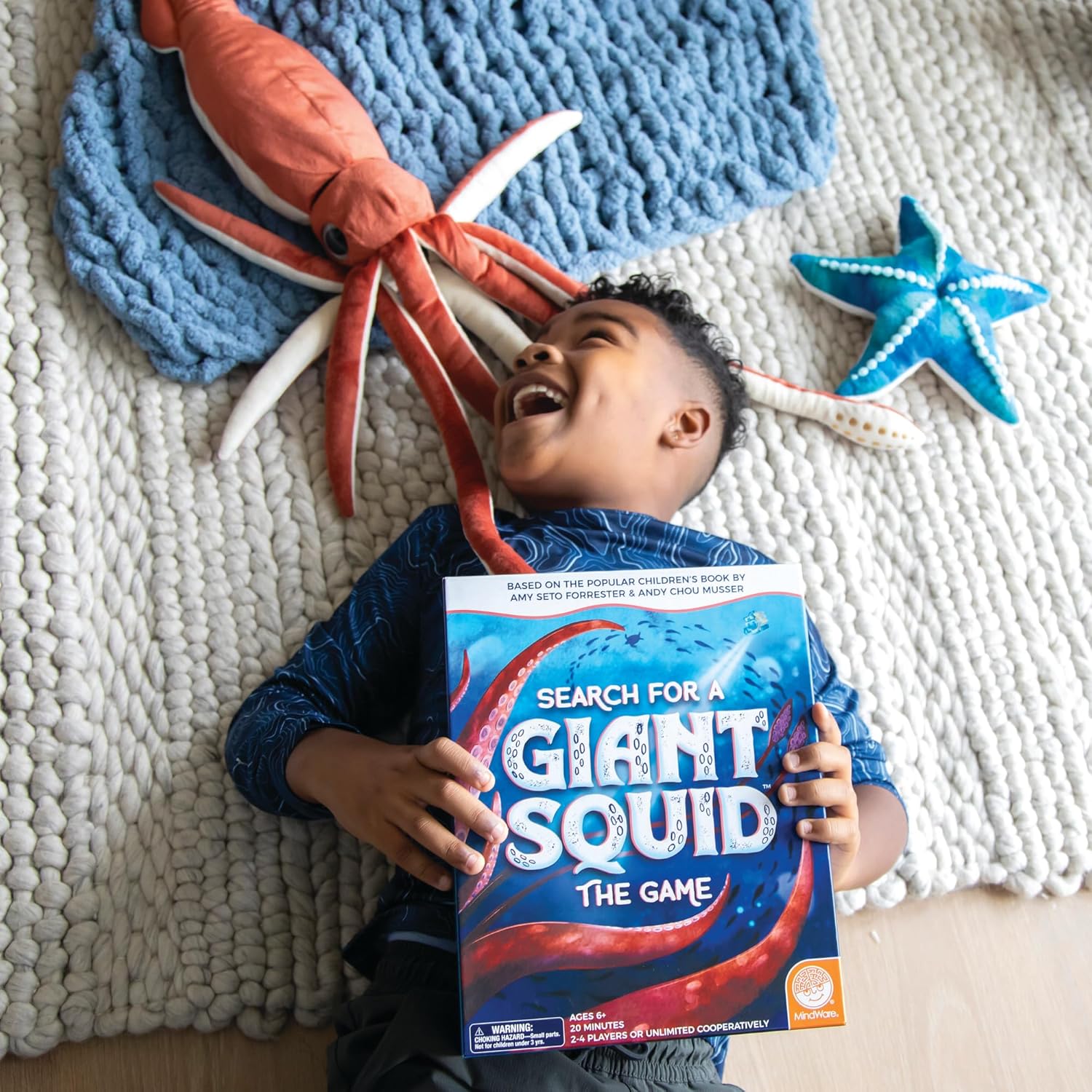 Joyful boy laughing, holding "Search for a Giant Squid" board game, surrounded by plush squid and starfish