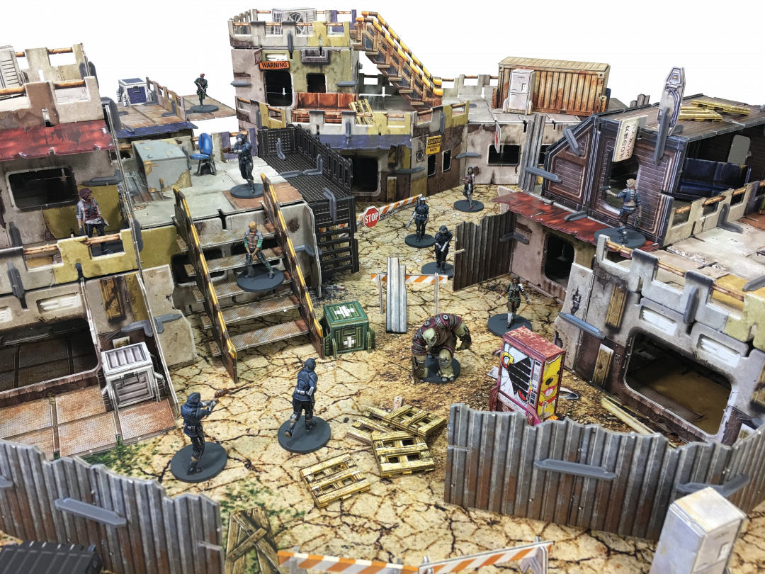 Tabletop game: Armed humanoid and creature miniatures deployed across multi-level, dilapidated buildings and fences in a sci-fi wasteland.