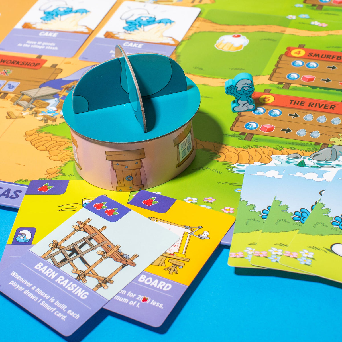 A colourful Smurfs board game featuring a 3D mushroom house, a blue Smurf meeple on &quot;The River&quot; path, and various game cards.