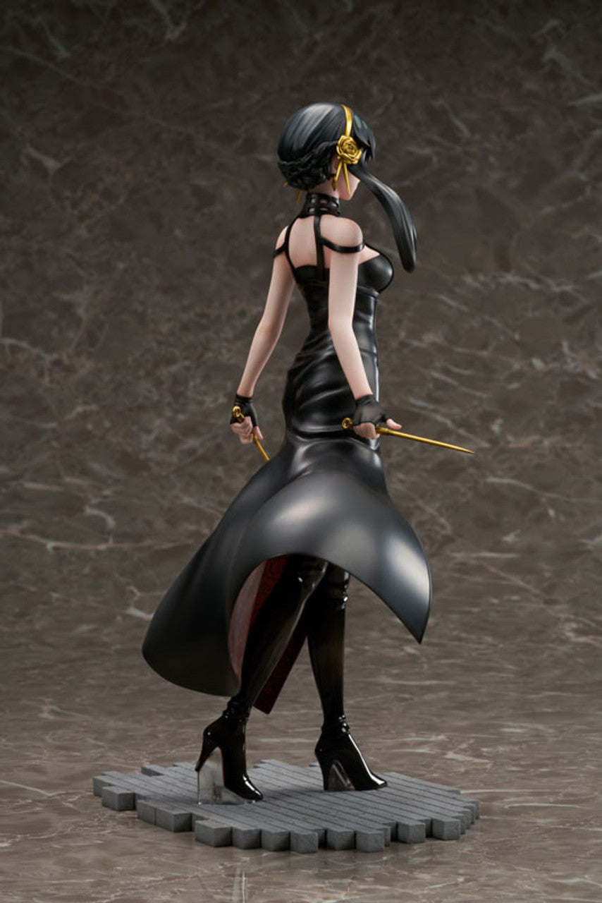 A Spy X Family Yor Forger figure in a black dress and boots, holding two gold weapons, with dark hair and a gold rose accessory, standing on a grey base
