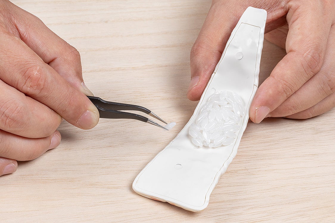 Hands use tweezers to place a rice grain into a white mold filled with other grains