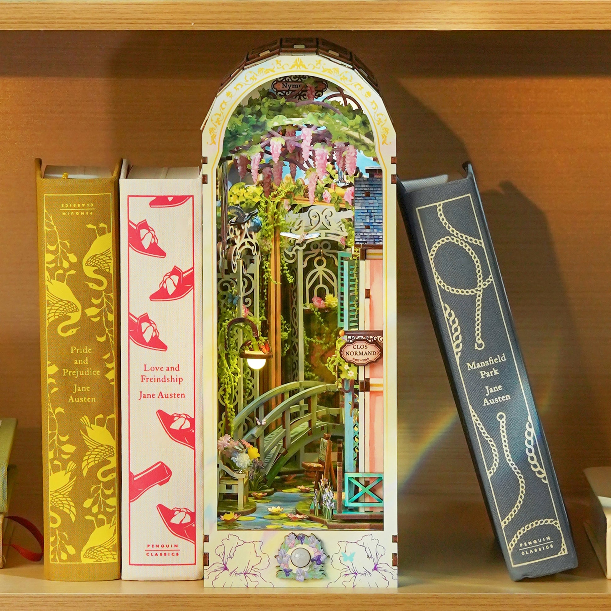 Miniature garden book nook with a bridge and wisteria, illuminated and placed between Jane Austen novels on a shelf