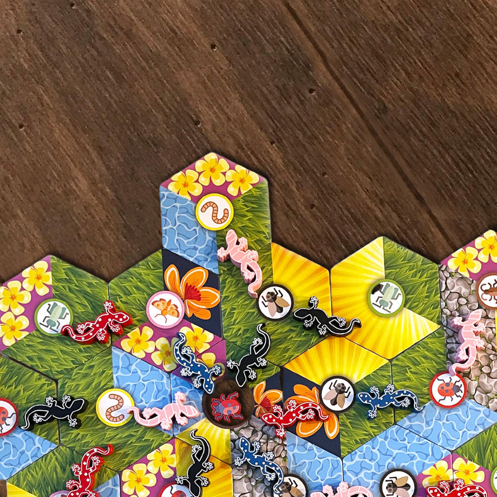 Overhead shot of a vibrant board game setup with hexagonal tiles depicting terrains and numerous colorful lizard and insect pieces