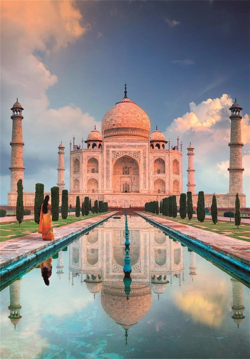 Woman in orange sari gazes at the majestic Taj Mahal, perfectly reflected in a long pool under a cloudy sky