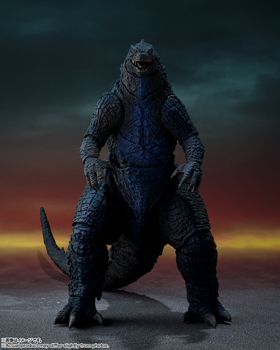 A dark blue Godzilla figure with textured skin and open mouth stands powerfully against a dramatic, fiery sky.