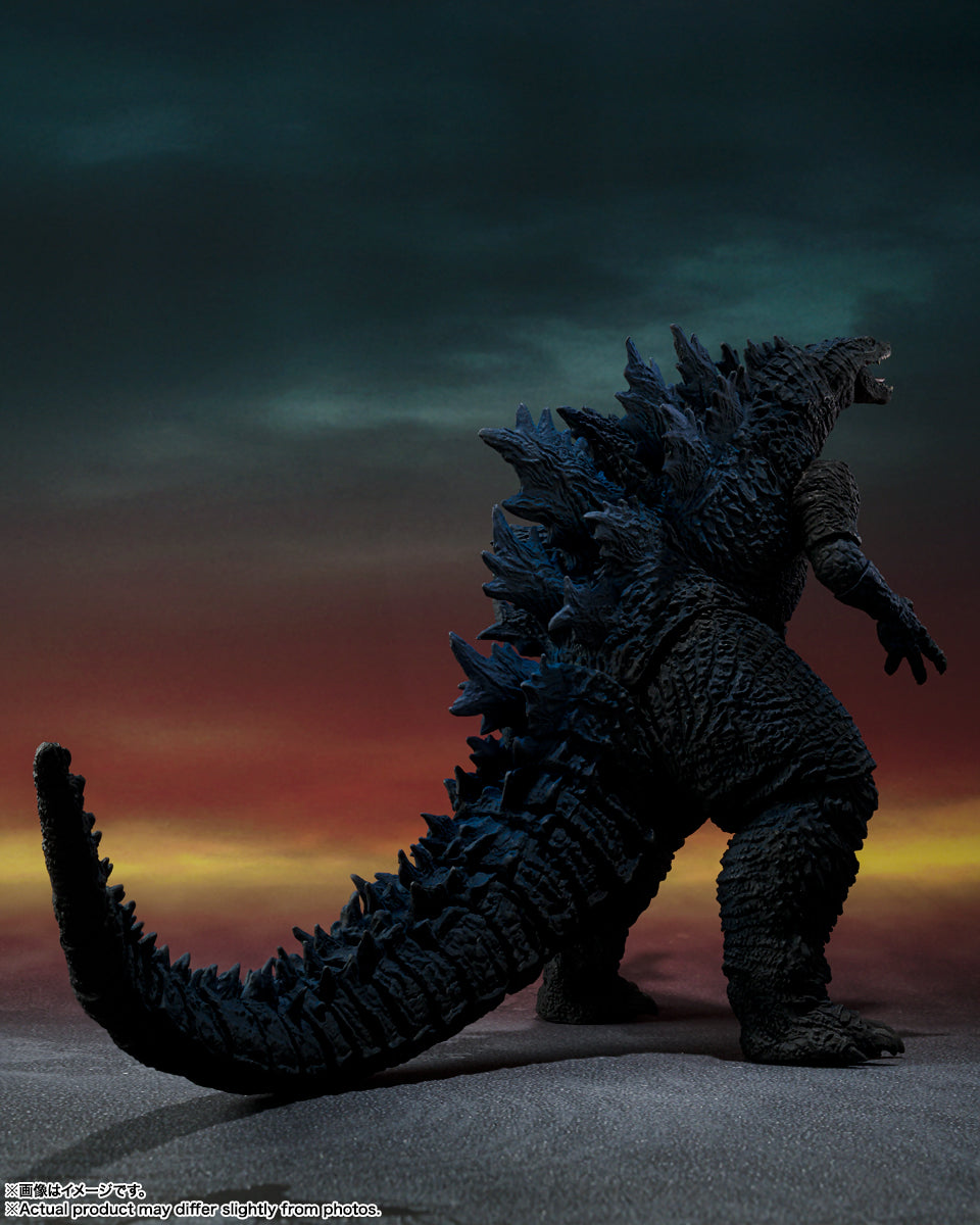 Godzilla stands menacingly, facing right, with its mouth open and prominent blue dorsal fins, against a dramatic sunset.