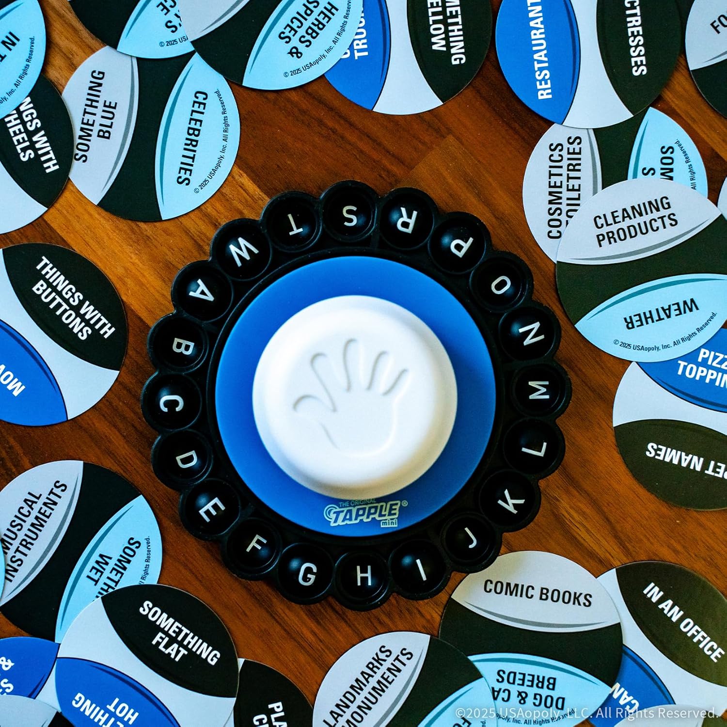 An overhead view of a Tapple Mini game board with a central white hand icon button and letter keys, surrounded by category cards on a wooden table