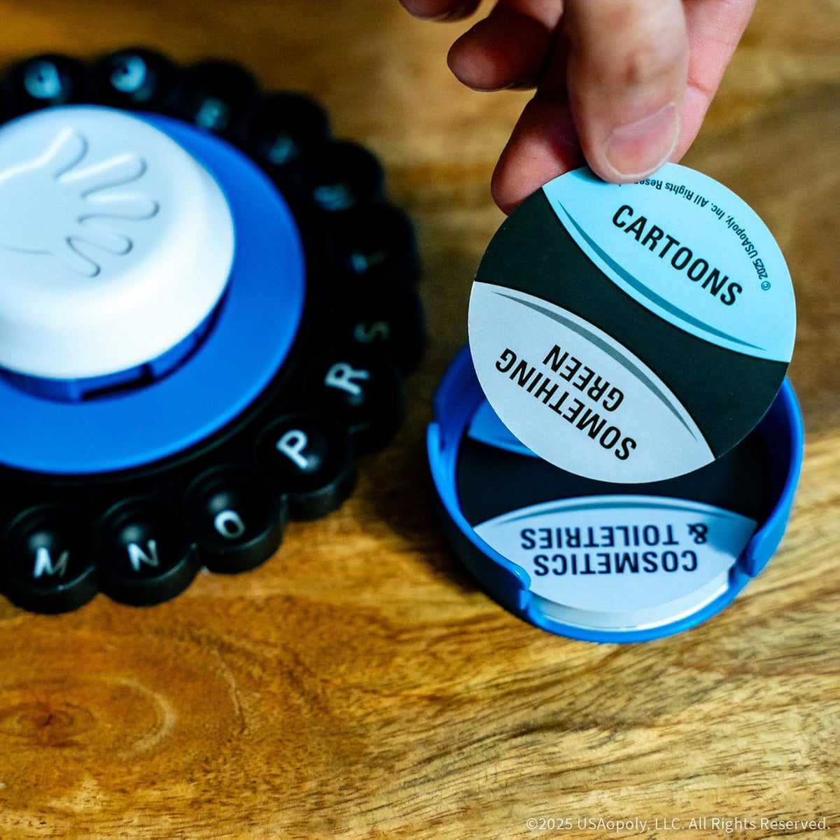 A hand holds a &quot;CARTOONS&quot; game card over a blue holder. Part of a letter spinner and a wooden table are visible
