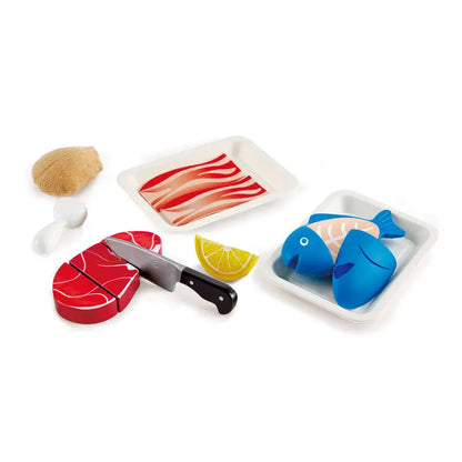 Child's play food set featuring a toy knife cutting a red steak. Also includes blue fish, lemon, brown coconut, and bacon tray.