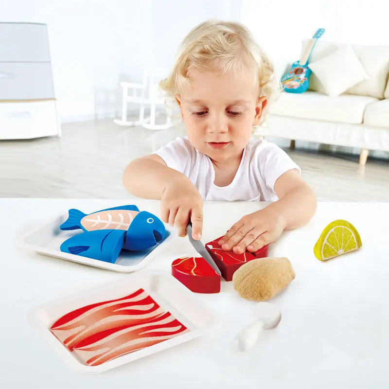 A young child uses a toy knife to cut a red block of play food meat, with toy fish and other food on the table.