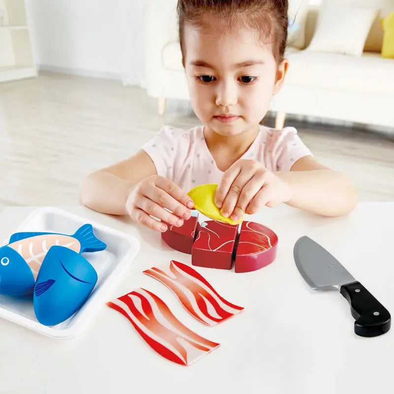 A young girl playing with a wooden toy food set, holding a lemon piece over a red steak. Toy fish, bacon, and a knife are on the table.