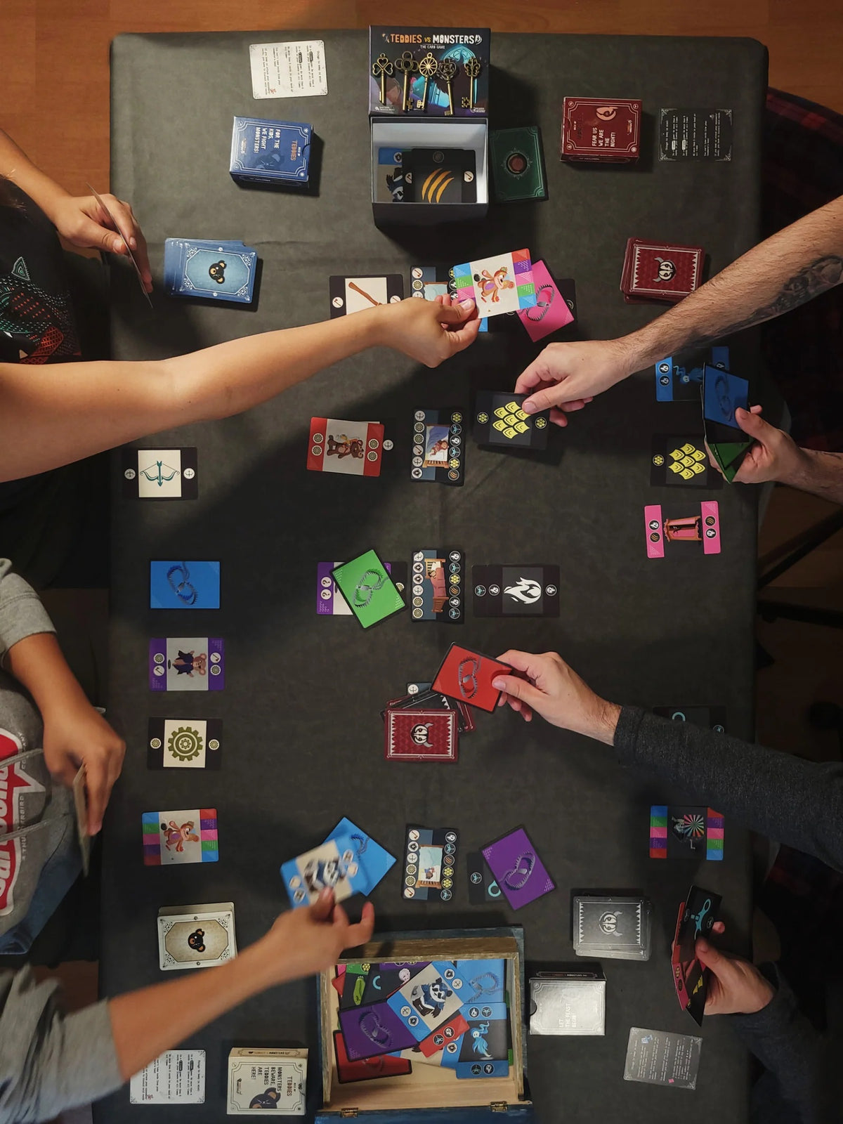 Overhead view of several hands playing the &quot;TeddieS vs MonsterS&quot; card game on a dark table with many cards