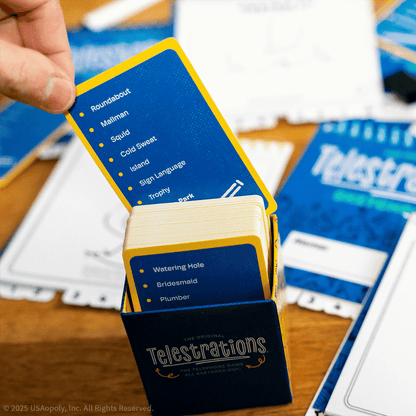 A hand draws a blue and yellow Telestrations game card from a deck, revealing words like 'Roundabout' and 'Mailman'