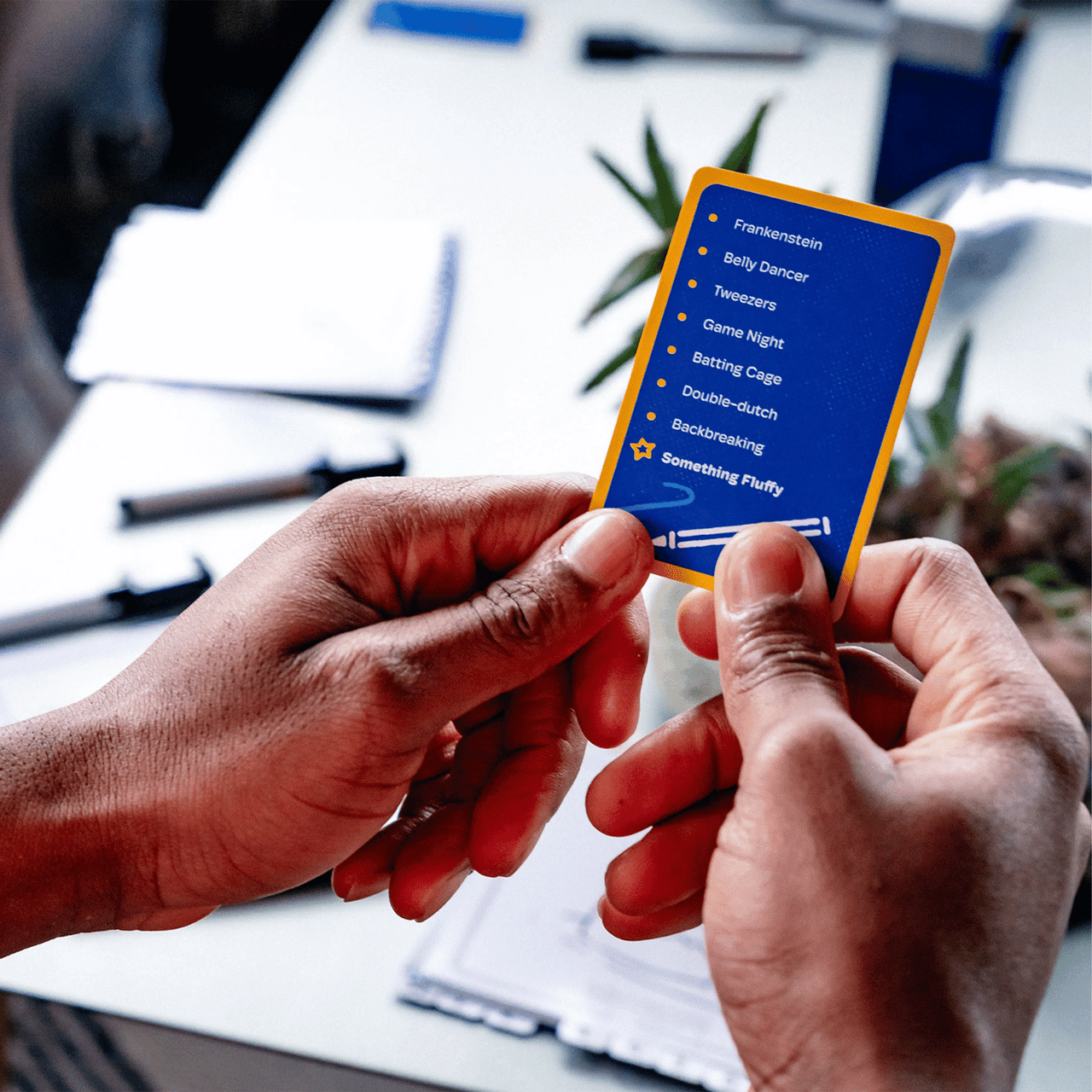 Two dark-skinned hands hold a blue game card with a yellow border, showing words for a game, like 'Frankenstein' and 'Something Fluffy'