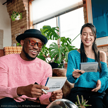 Man and woman laughing, playing a "Sketch It!" game with small drawing pads