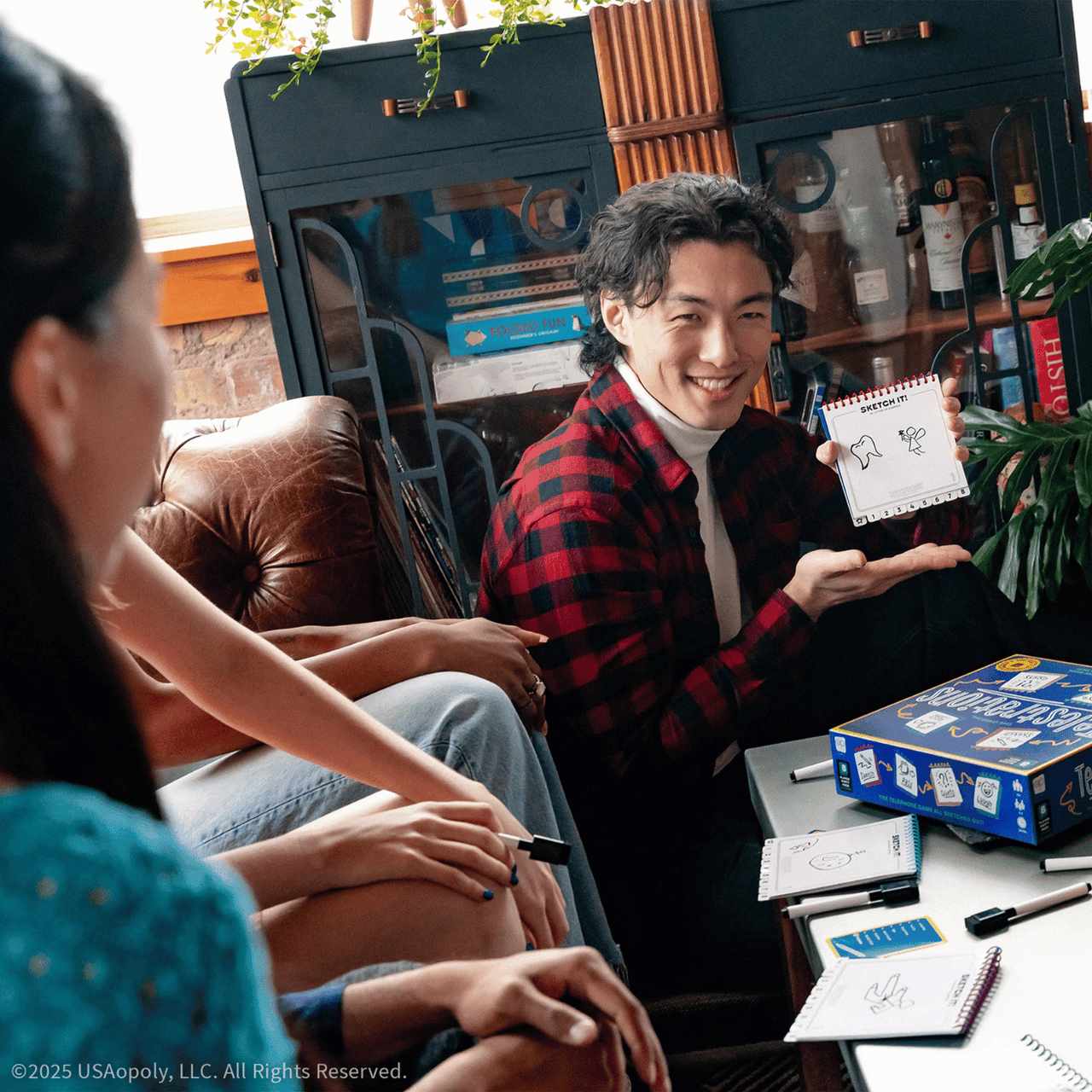 Smiling man in a plaid shirt holds up a sketch, playing Telestrations board game with friends