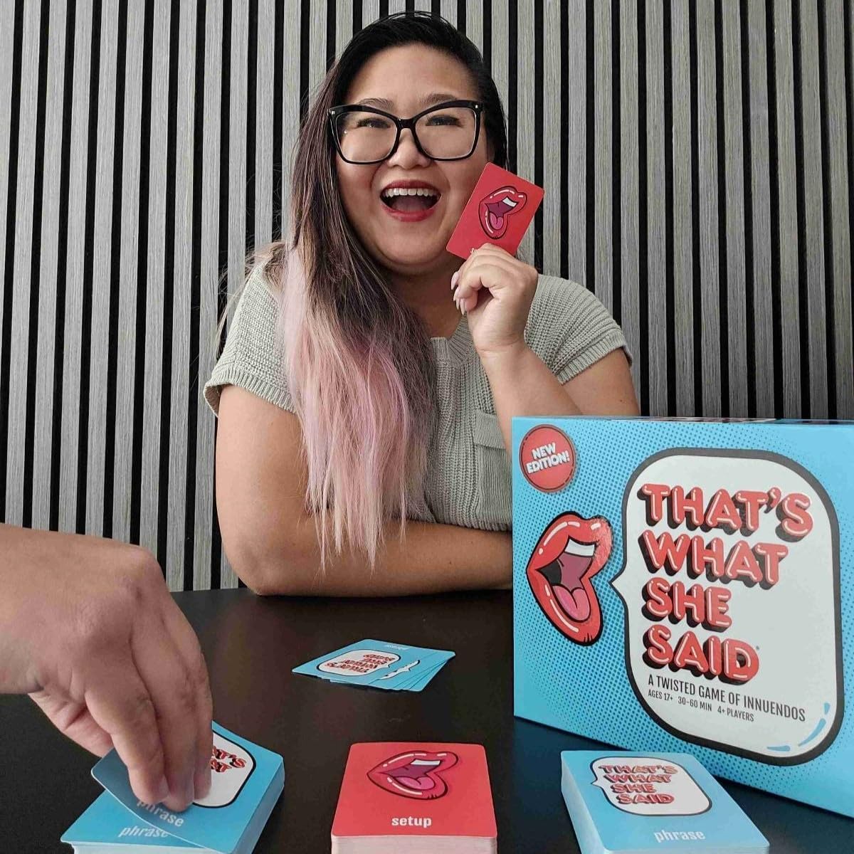 A woman with pink hair and glasses laughs, holding a red card from the "That's What She Said" board game