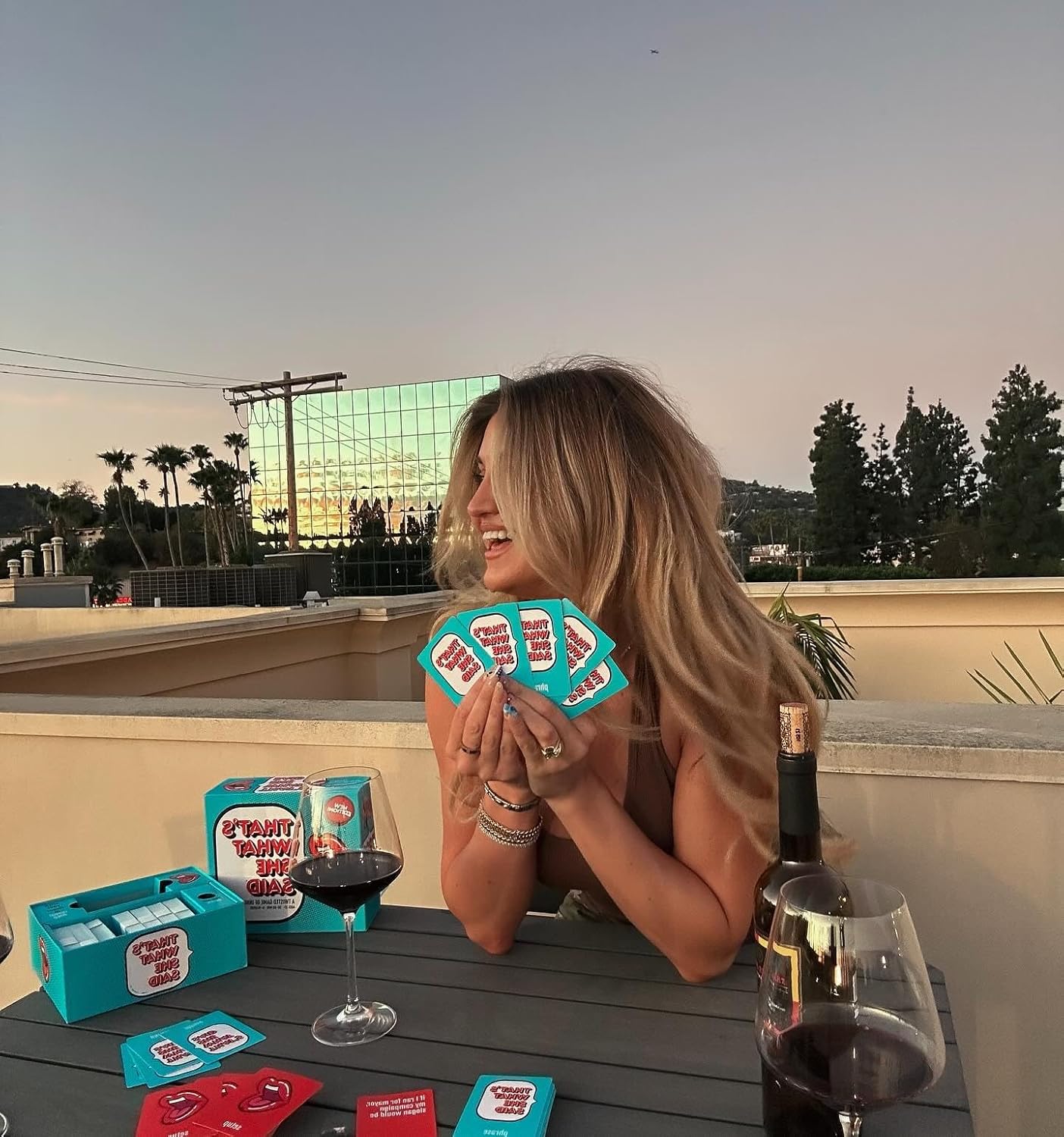 Smiling woman laughs, holding aqua "That's What She Said" cards while playing on a rooftop with wine at sunset