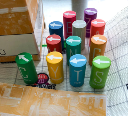 Colourful cylindrical game pieces, each with an arrow and a Tokyo Metro line name, are scattered on a white grid-patterned surface