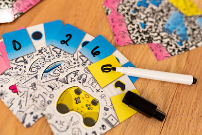 Dry-erase marker writing "6" on a yellow game card, surrounded by blue number and doodle art cards