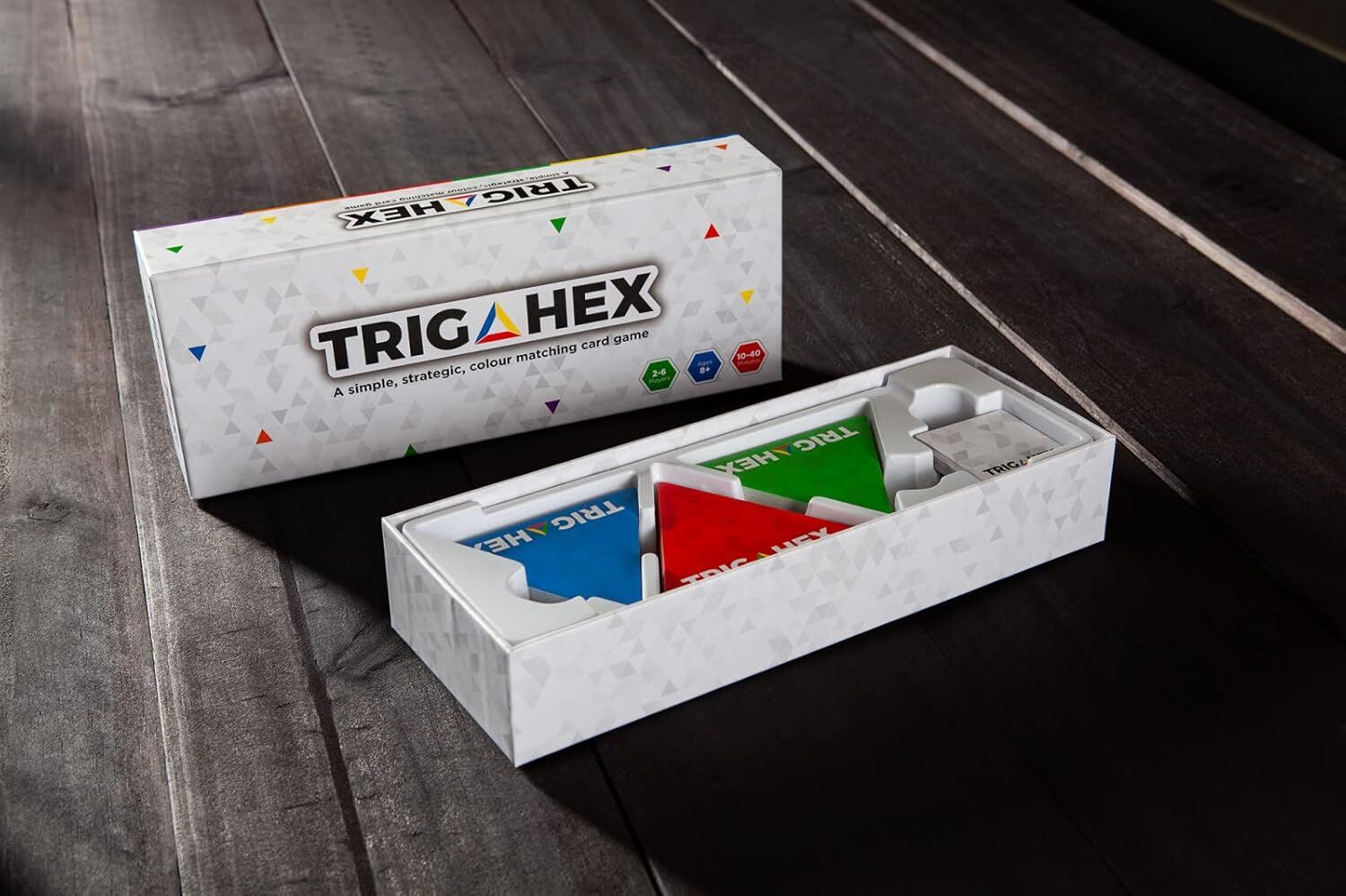 Open TRIGA HEX game box shows blue, red, green triangular components, with a closed box behind it on wood