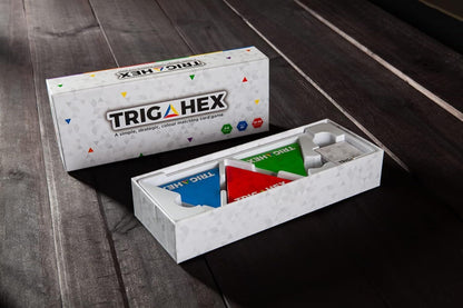 Open TRIGA HEX game box shows blue, red, green triangular components, with a closed box behind it on wood