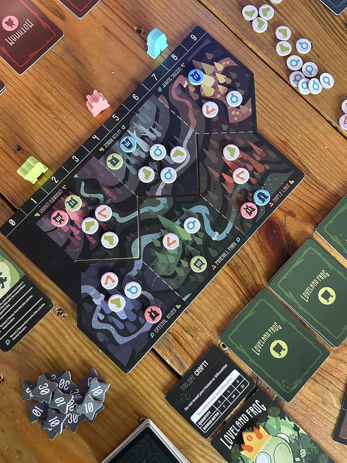 A dark game board, player pieces, tokens, and cards from the Twisted Cryptids Cryptid Culture Expansion game are laid out on a wooden table