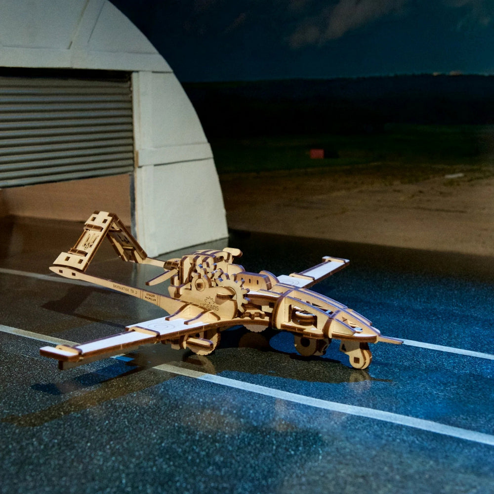 Detailed wooden Bayraktar TB2 drone model with Ukrainian trident symbol, resting on an airfield tarmac near a hangar
