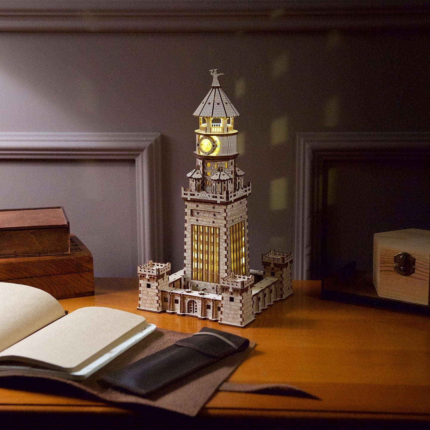 A glowing, intricately detailed wooden tower model sits on a dark desk, next to an open journal
