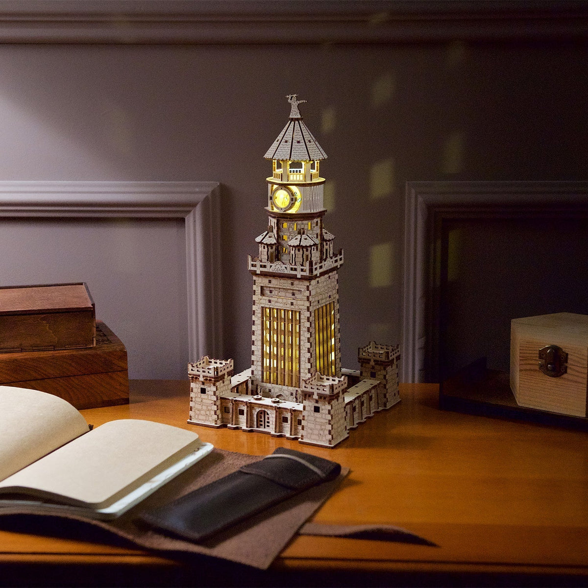 A glowing, intricately detailed wooden tower model sits on a dark desk, next to an open journal