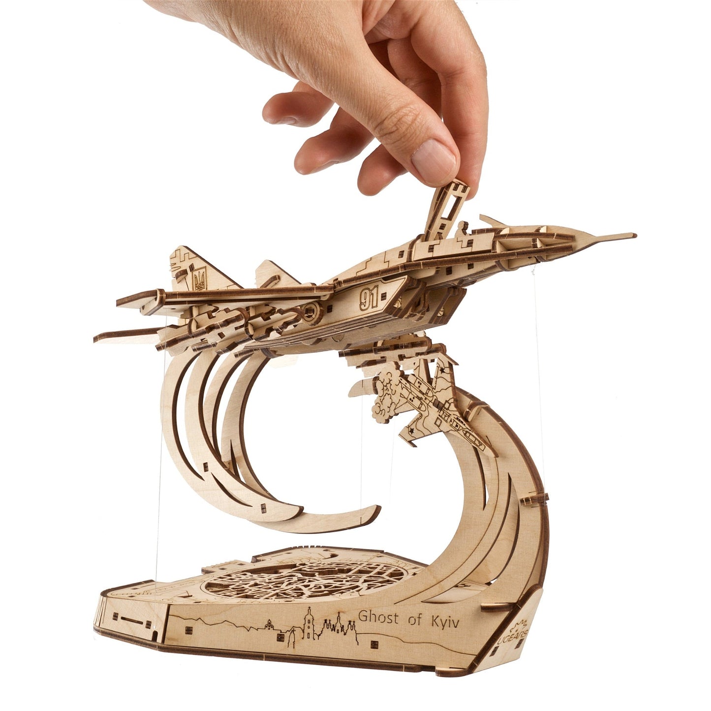 Hand adjusts the canopy of a wooden MiG-29 fighter jet model, with Ukrainian symbols and "Ghost of Kyiv" stand