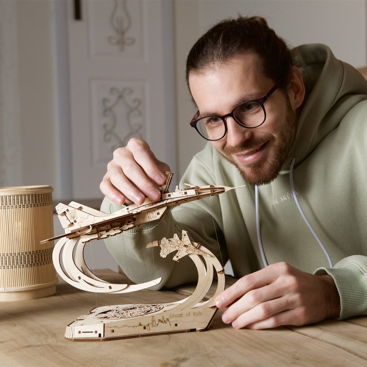 Smiling man assembling a wooden fighter jet model with a "Ghost of Kyiv" stand