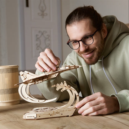 Smiling man assembling a wooden fighter jet model with a "Ghost of Kyiv" stand