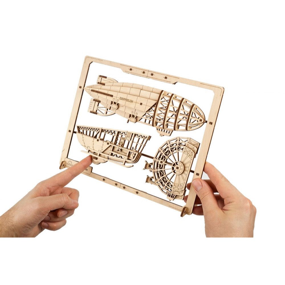 Hands hold a wooden Zeppelin airship model kit with "ZEPPELIN" etched on parts, still attached to its frame
