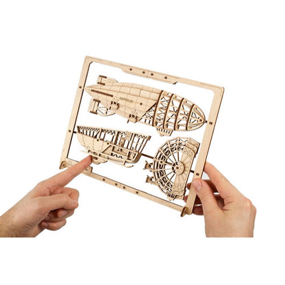 Hands hold a wooden Zeppelin airship model kit with "ZEPPELIN" etched on parts, still attached to its frame