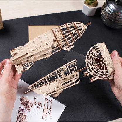 Two hands assemble a laser-cut wooden zeppelin model, connecting parts over instructions on a desk