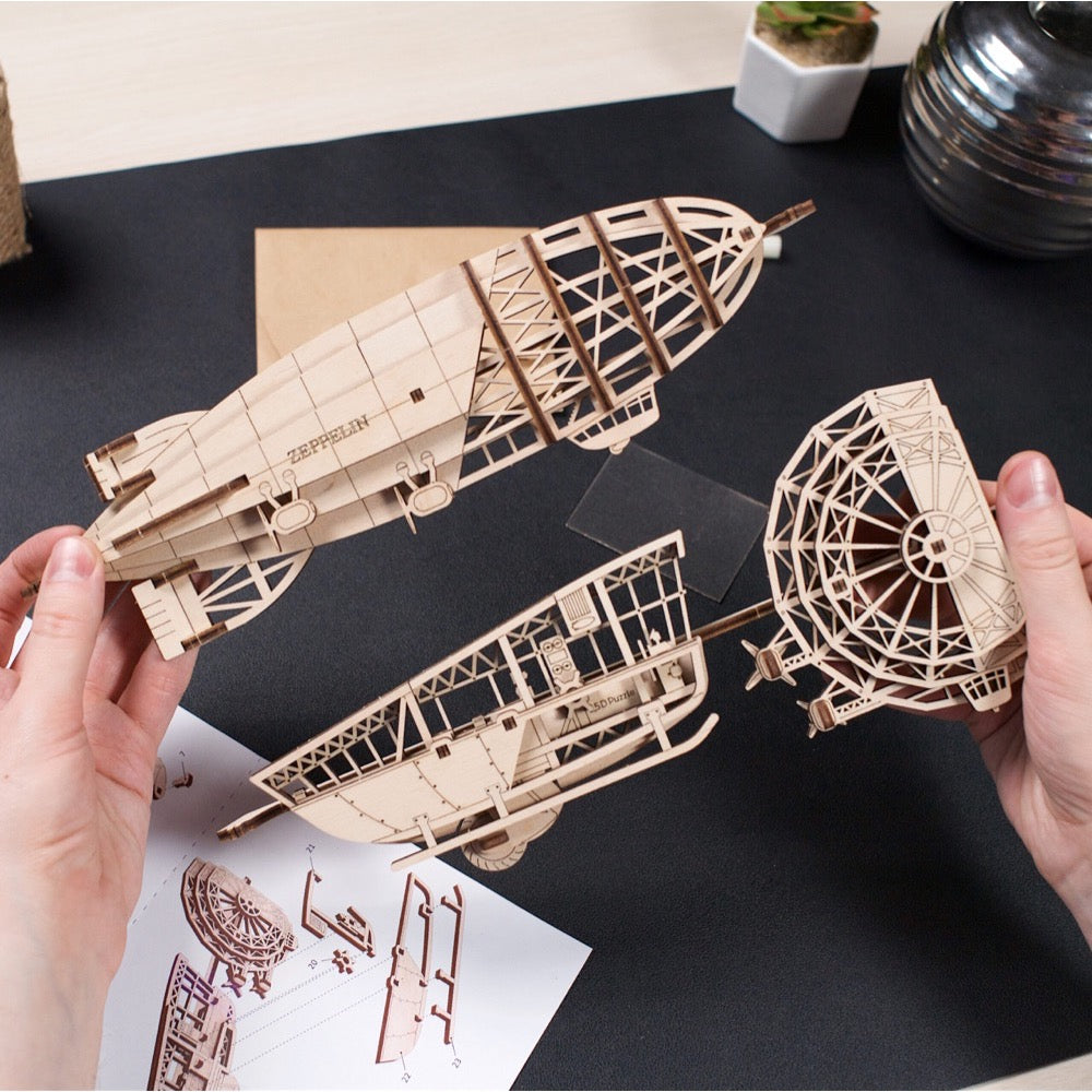 Two hands assemble a laser-cut wooden zeppelin model, connecting parts over instructions on a desk