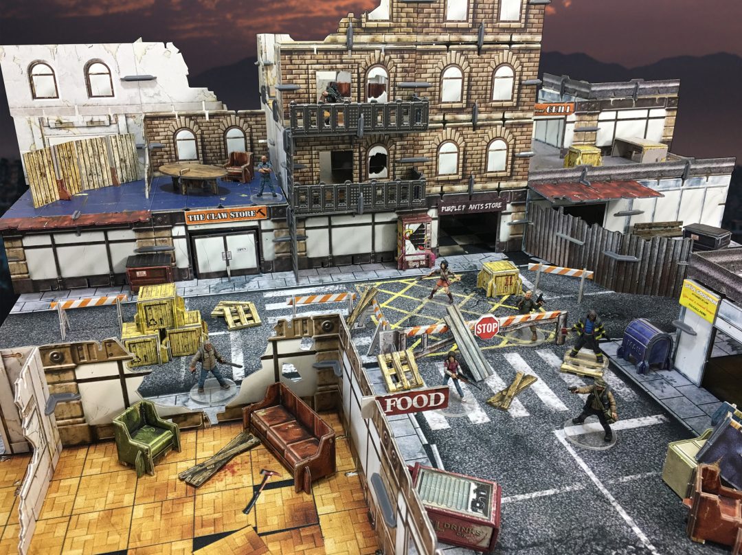 A tabletop wargame of a ruined city. Miniature figures stand among damaged buildings, street barricades, and debris under a dark sky.