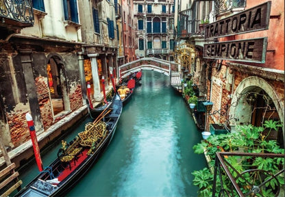 Two ornate gondolas in a narrow Venice canal flanked by historic buildings, an arch bridge, and a "Trattoria" sign.