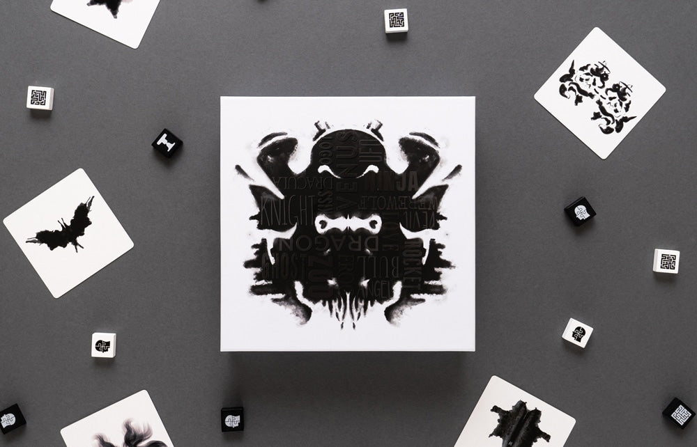 Rorschach-style inkblot game box with playing cards and dice on a gray background