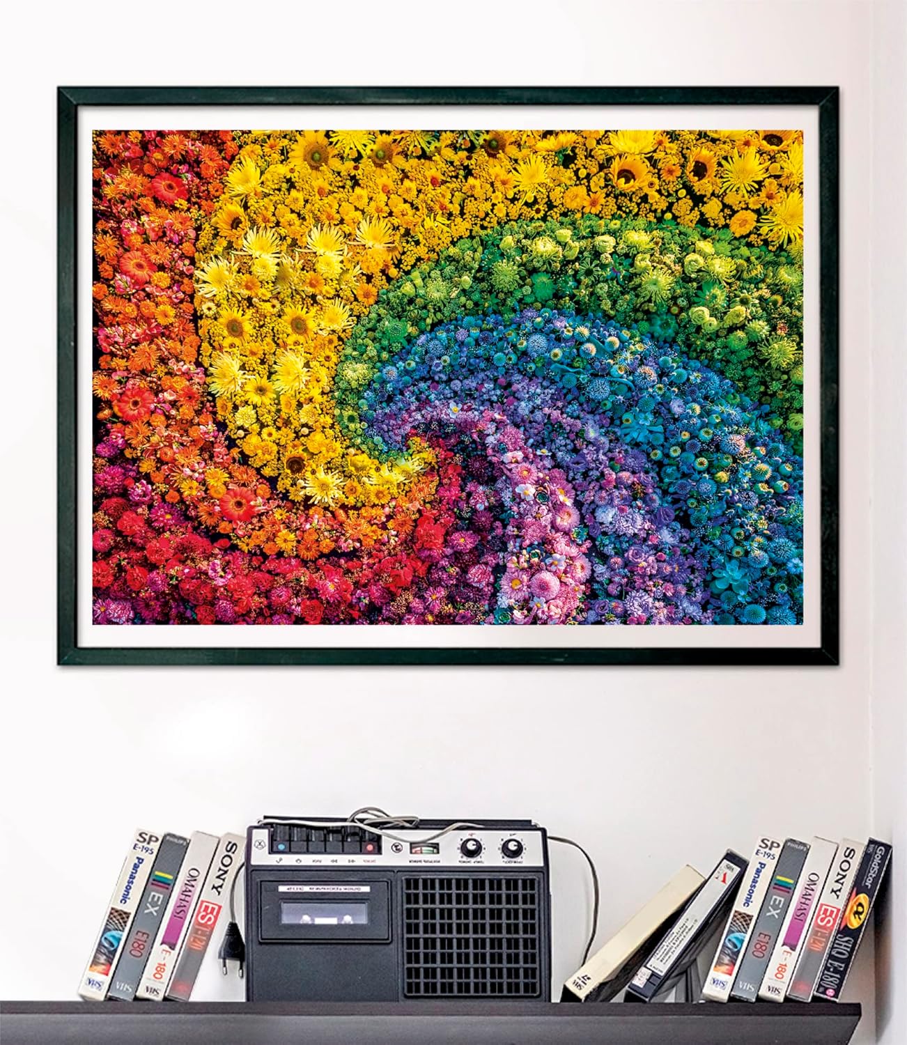 A framed artwork of a vibrant rainbow spiral made of tiny flowers hangs above a shelf with a cassette player and VHS tapes
