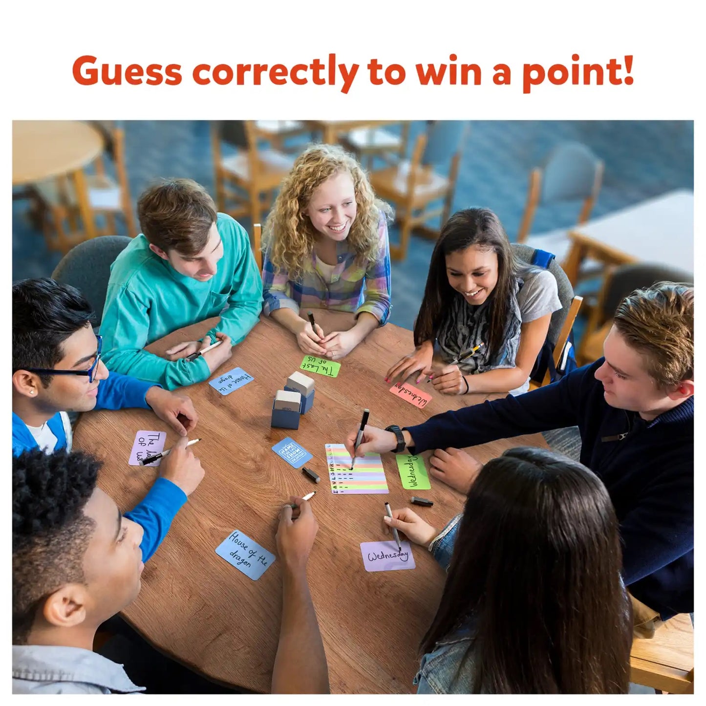Six students happily playing a writing and guessing card game around a wooden table