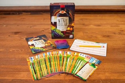The Wine Cellar board game box, rulebook, client cards, score pad with a pencil, and a fan of colorful game cards are displayed on a wooden surface