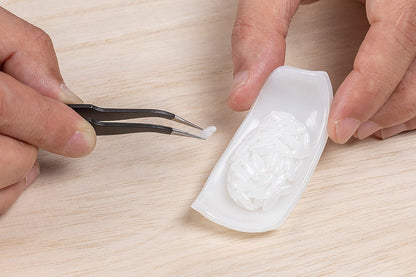 Hand holding tweezers picks up a white, rice-shaped pellet from a small tray of similar pellets on wood
