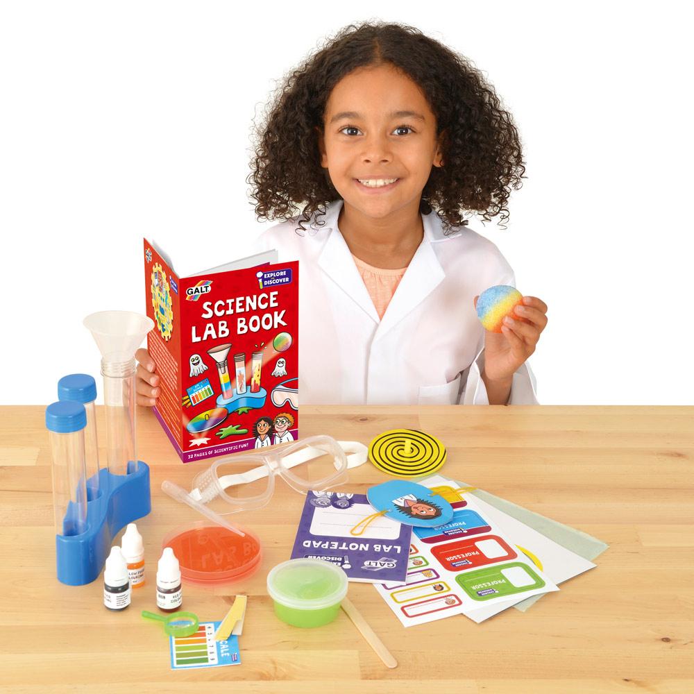 Smiling girl in a lab coat with a science lab kit, including a book, test tubes, and safety glasses